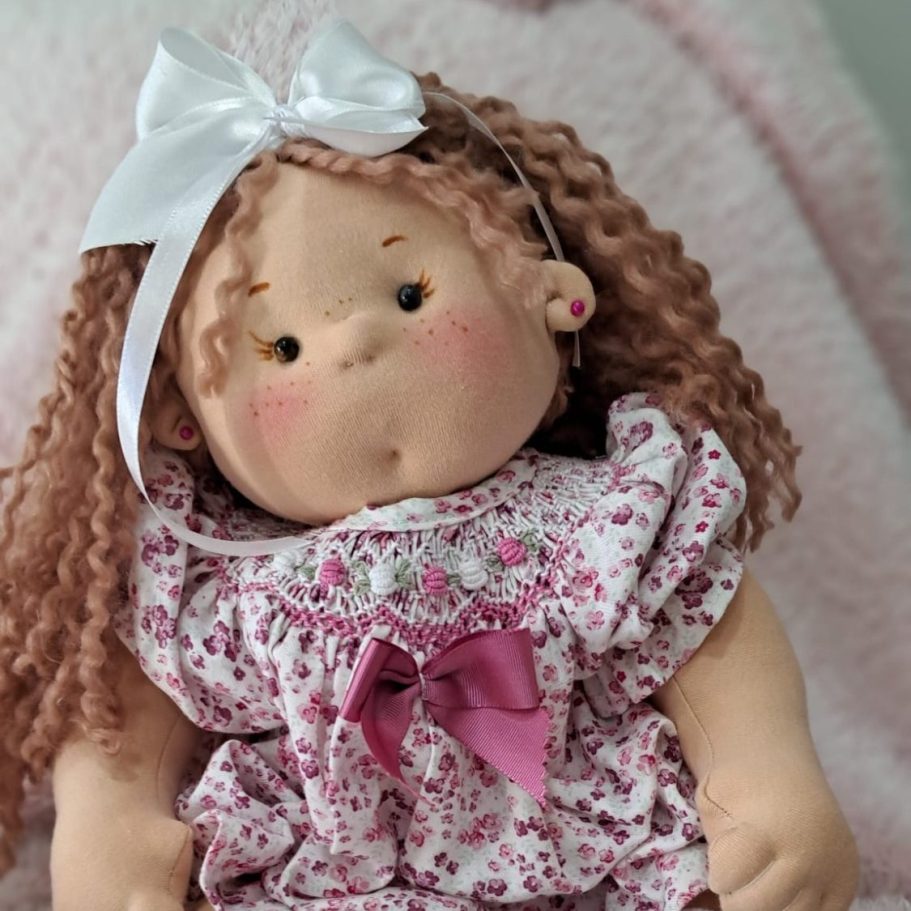 Muñeca de tela con pelo rizado, vestida con un vestido floral y un lazo rosa.