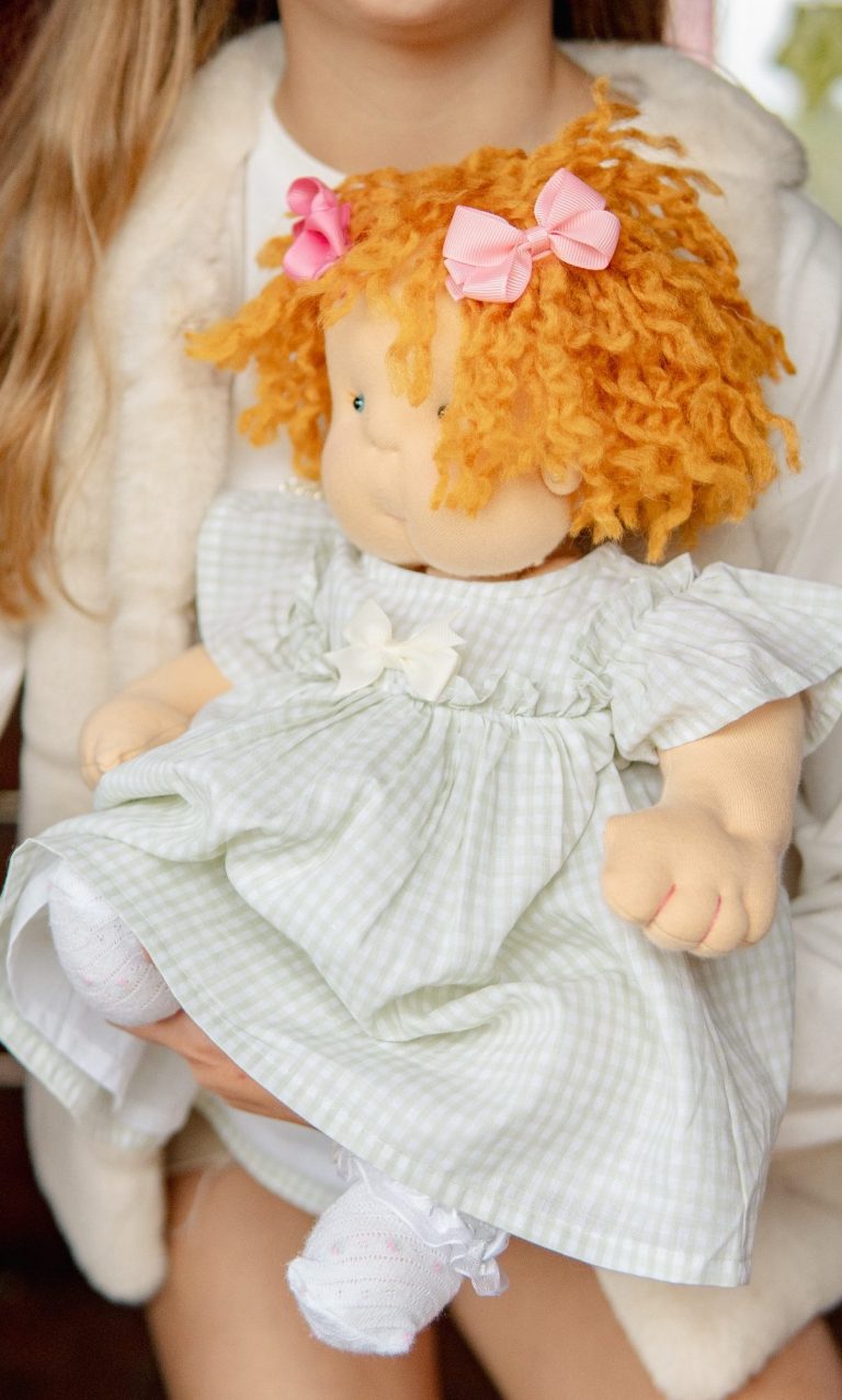 Niña con cabello rizado y muñeca vestida con un vestido claro y cintas rosas.