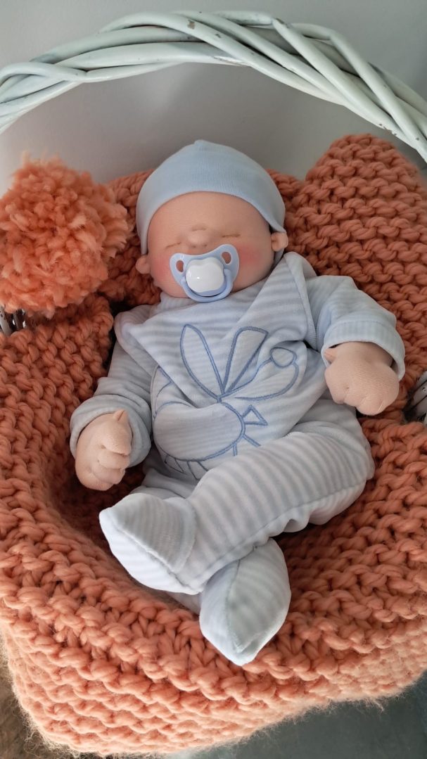 Bebé de tela con gorro azul y chupete, pijama de rayas azules, sobre una manta de punto terracota