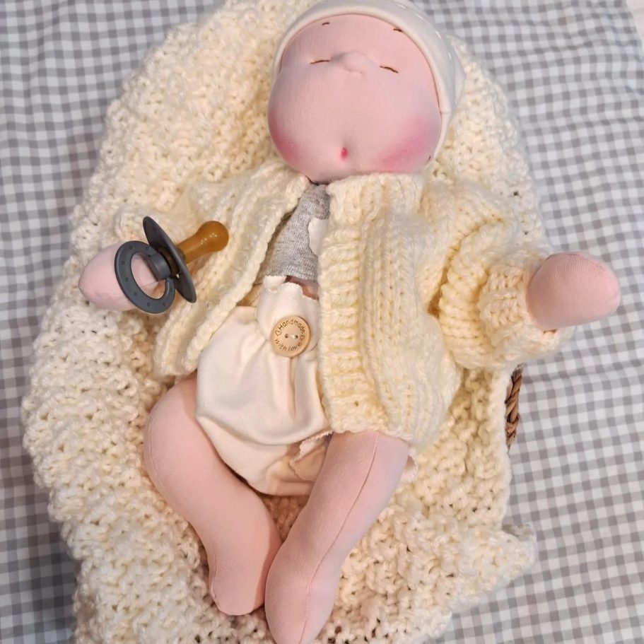 Muñeca de trapo con chupete, vestida con un suéter amarillo y envuelta en una manta.